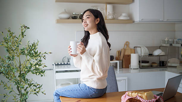 istock Young woman holding a coffee mug while sitting on a kitchen table, looking away with a thoughtful smile. 2227698024