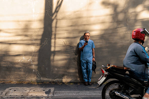 Portrait of a senior healthcare professional in a blue uniform and eyeglasses, leaning casually against a wall on the street. He holds a bag in one hand and a phone in the other, while a motorcyclist passes by in the background, adding a dynamic element to the everyday urban scene.