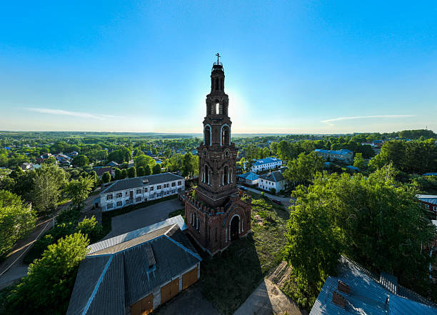 An aerial shot captures a tall, historic bell tower in Yuryev Polsky, Russia, prominently illuminated by direct sunlight, with the town spreading out behind it under a clear sky