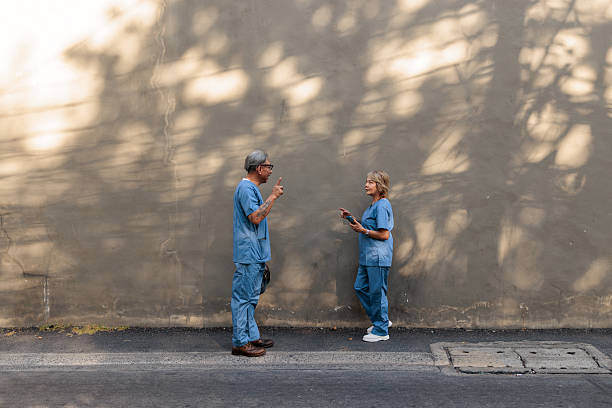 A senior male and female nurse in their uniforms, meet on the street and engage in conversation. The woman holds her phone in her hands as they share a friendly moment during their busy day outside the hospital.