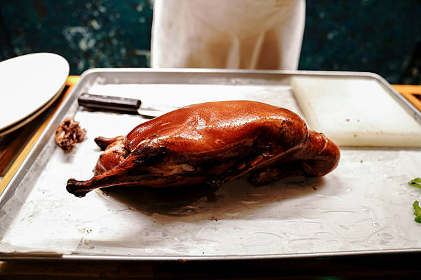 A close-up shot of a chef carving a whole roasted duck with a large cleaver on a cutting board, wearing protective gloves. The golden-brown crispy skin of the duck glistens under warm kitchen lighting, highlighting its texture and succulence. This image captures the essence of traditional culinary preparation and fine dining presentation.