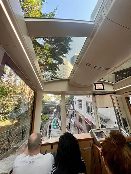 Hong Kong, China - January 03, 2025: View from inside a Peak Tram carriage in Hong Kong, as it ascends toward Victoria Peak. The modern funicular carriage features large panoramic windows and a glass roof, offering passengers a steep-angled perspective of the surrounding trees and city skyline. Passengers can be seen seated in the foreground, looking out toward the tracks and a bridge ahead