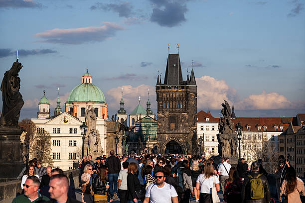 Tourists and locals stroll across Charles Bridge in Prague during golden hour, surrounded by statues and historic architecture.
