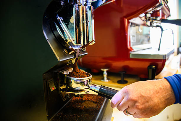 A close-up of a barista preparing freshly ground coffee using a commercial espresso grinder. The coffee grounds fall into a portafilter in front of a vibrant red espresso machine, capturing the process of artisan coffee preparation.