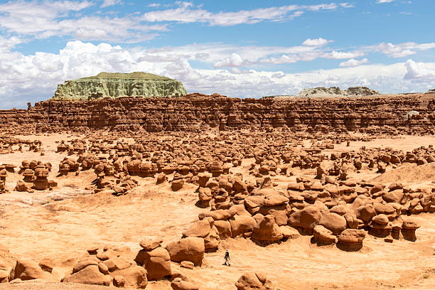 One person walkin between mushroomlike sandstone rock formations called goblins and eroded cliffs in Goblin Valley State Park, Utah, USA against a white clouded blue sky