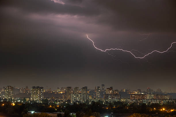 A powerful lightning bolt flashes across the dark, stormy sky during a summer thunderstorm at night. Below, the illuminated skyline of Kyiv, Ukraine, glows in the darkness