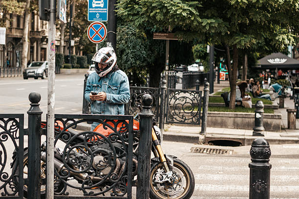 A man wearing a helmet and denim jacket stands next to his parked motorcycle while checking his smartphone on Rustaveli Avenue in Tbilisi, Georgia. The urban setting includes decorative iron fencing, street signs, and pedestrians sitting in a shaded public area. The image reflects everyday city life, modern transportation, and digital connectivity in a vibrant urban environment.