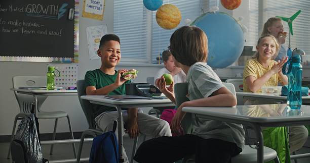 istock Elementary School Student Sitting at Desk, Eating Homemade Sandwich from Lunch Box, Talking to Classmate 2227773225