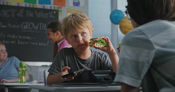istock Elementary School Student Eating Sandwich from Lunch Box, Using Phone and Talking to Classmate 2227773451