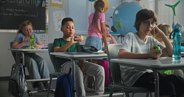 istock Elementary School Student Sitting at Desk, Eating Homemade Sandwich from Lunch Box, Talking to Classmate 2227775249