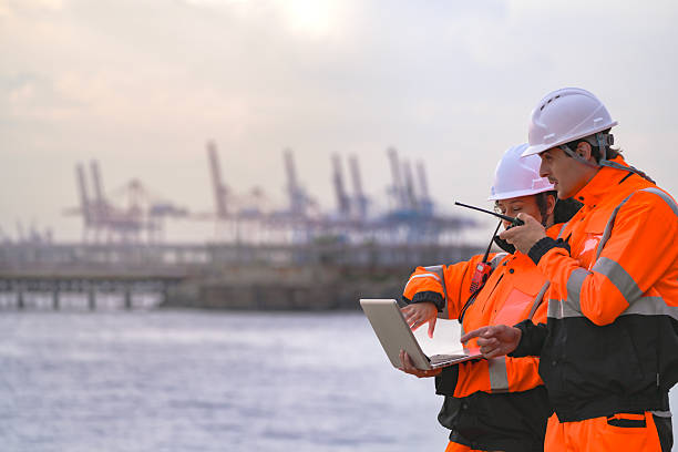 istock Two engineers wearing safety uniforms and hard hats communicate using a radio while reviewing data on a laptop by the sea during sunset. Maritime industrial setting. 2227784861