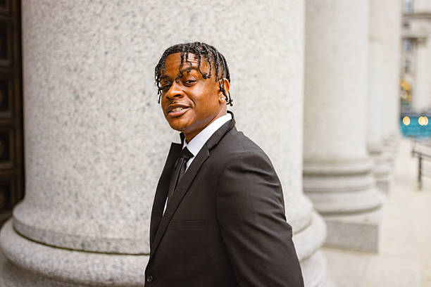 A confident young man in formal attire standing in front of a classical urban architectural structure, looking towards the camera.