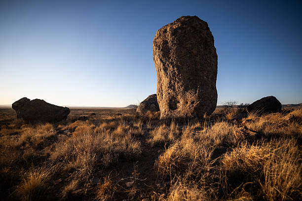 A massive solitary rock formation stands in dry desert grasslands, lit by the setting sun with a wide-open sky above.