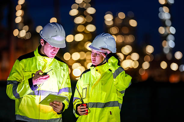 istock Two engineers in high-visibility jackets and safety helmets work during the night shift at an industrial facility, reviewing data on a tablet and coordinating via walkie-talkie. 2227785589