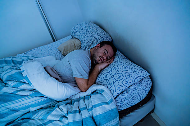 A man with Down syndrome lies comfortably in bed, wearing a light blue t-shirt. He appears peaceful and relaxed on a bed with blue and white striped bedding and decorative patterned pillows. The soft, dim lighting creates a calm, restful atmosphere in the bedroom.