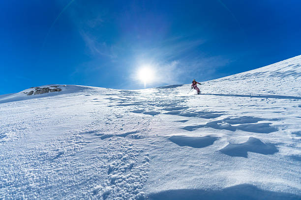 A male ski tourer dressed in bright attire descends a pristine snowy mountain under a clear blue sky with bright sunlight.