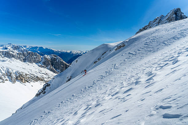 A male ski tourer expertly descends a pristine snowy mountain slope on a sunny day, surrounded by breathtaking alpine landscapes under a vibrant blue sky.