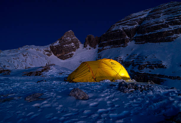 A glowing yellow tent rests on a snowy mountain slope under a dramatic alpine sky during sunrise. Rugged peaks surround the serene campsite.