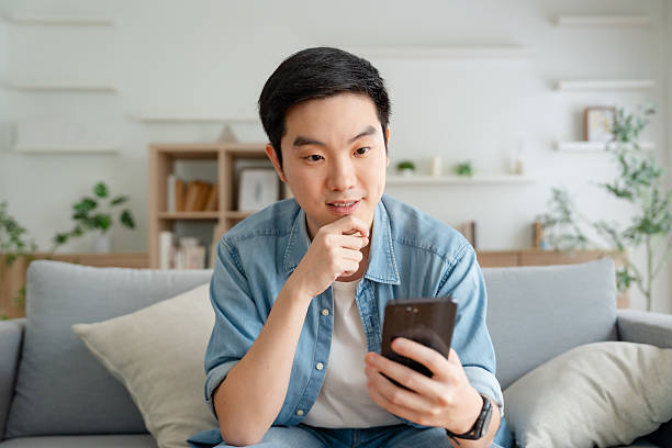 istock Smiling handsome young Asian man sitting on cozy couch, using funny mobile apps in living room. man at home, doing online shopping, messaging friends, posting on social media 2227794020