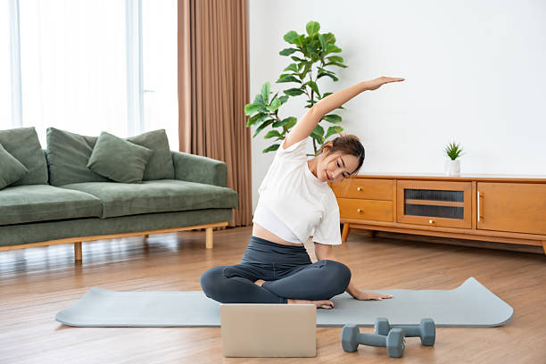 istock Young Asian woman stretching yoga workout on exercise mat while online training class with computer laptop in living room 2227794153
