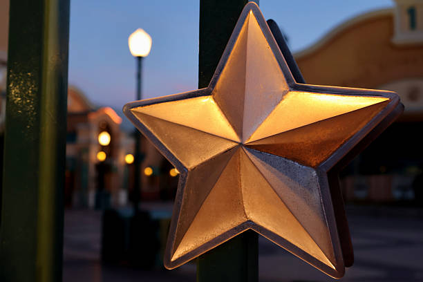 An illuminated metallic star sculpture stands prominently in a thematic amusement park, bathed in the glow of evening lights. This close-up view highlights the star's intricate design and polished surface, reflecting the vibrancy of the surrounding attractions. The scene blends playful creativity and a festive atmosphere, drawing visitors into a world of imagination and enchantment.