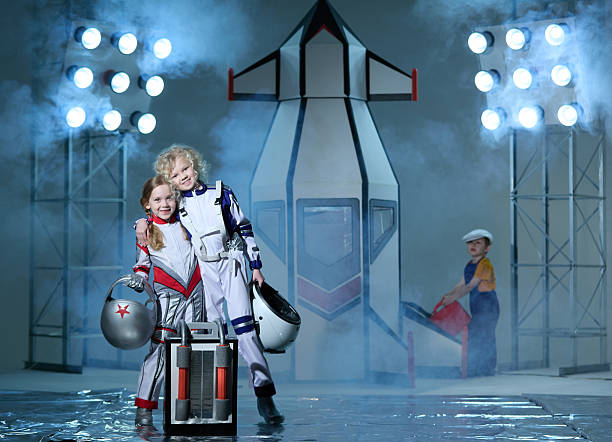 Two beautiful girls elementary age dressed in  astronaut costumes. The girls are standing in front of spaceship (rocket mockup from cardboard). Funny boy technical worker is fuelling rocket before a flight into space. Studio shooting