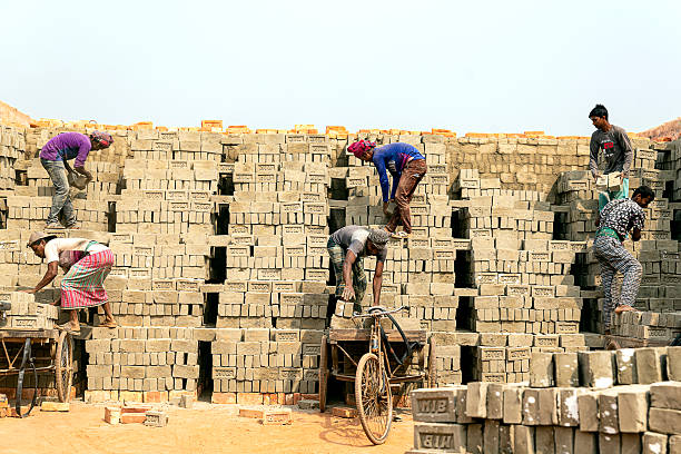 Manikganj, Bangladesh - 9 February, 2018: Laborers skillfully load and transport bricks on bicycles at a bustling brick kiln. Dust fills the air as they work diligently in the warm afternoon sunlight.