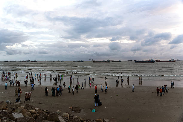 Cox's Bazar, Bangladesh - 1 July, 2019: Visitors stroll along a sandy beach during the evening. The shoreline is bustling with families and groups while cargo ships are anchored on the horizon under a moody sky.