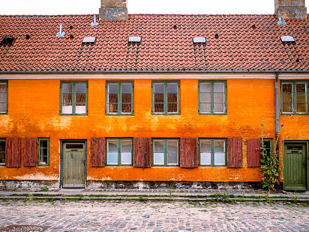 Historic orange row house with green windows and red-tiled roof in Nyboder, Copenhagen, Denmark; cobblestone street and climbing plants add charm to the scene.