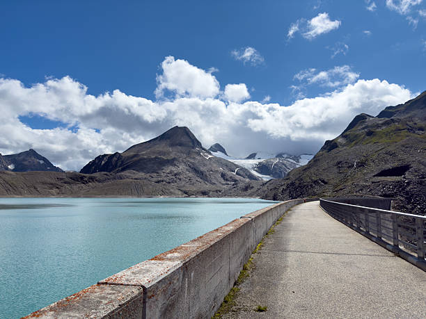 Calm turquoise mountain lake bordered by concrete dam under blue sky with clouds. Scenic alpine landscape with rocky peaks and glacier in background. Nature and outdoor travel destination.