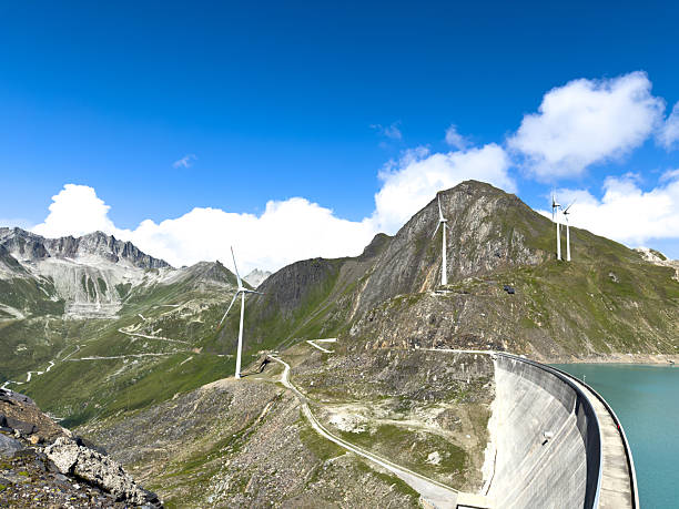 Large curved dam in mountainous terrain with wind turbines on ridges. Renewable energy infrastructure in alpine landscape with blue sky and scattered clouds. Sustainable power generation concept.