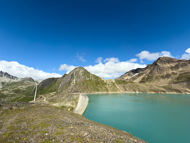 Turquoise mountain reservoir bordered by a curved dam and wind turbines on grassy hills. Scenic alpine landscape with clear blue sky and rugged peaks. Renewable energy and nature.