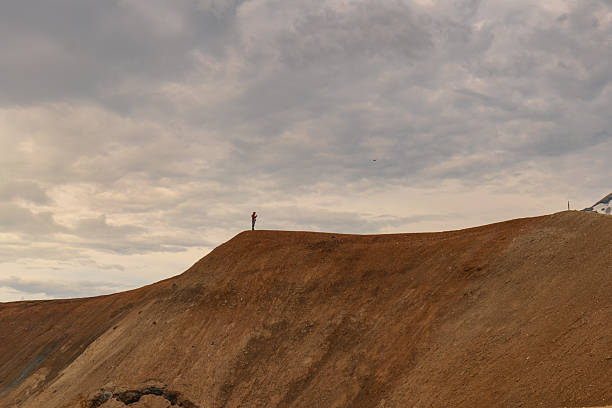 A person stands on a rust colored ridge under a cloudy sky, surrounded by barren, eroded slopes in a volcanic landscape typical of Iceland.