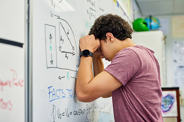 Confused student guy leaning on whiteboard looking stressed while solving complex equations. Tired high school teen struggling with math problem in classroom at secondary school. High school student showing frustration in front of science formula during lesson, attention deficit and ADHD.