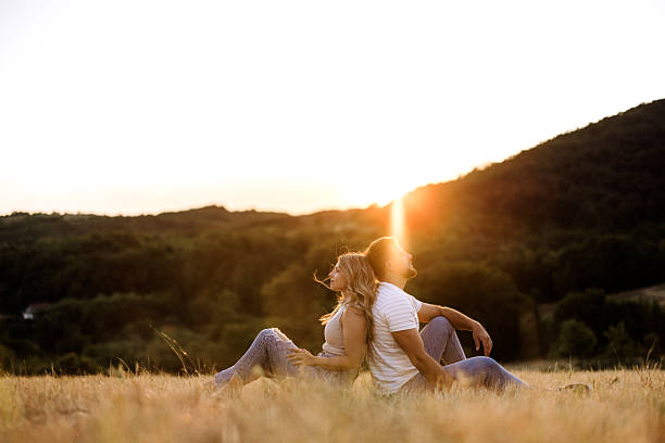 Young couple sitting on grass, back to back, in a peaceful meadow during golden hour. Surrounded by nature, they enjoy a quiet, intimate moment together, reflecting closeness and harmony.