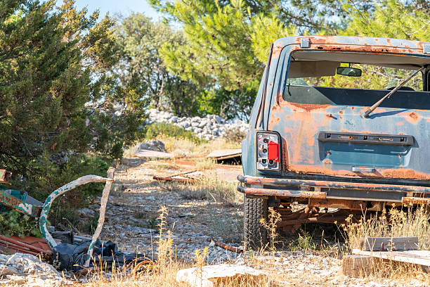Rear shot of a heavily corroded SUV with broken windows, surrounded by scattered construction debris and weeds in a remote area.