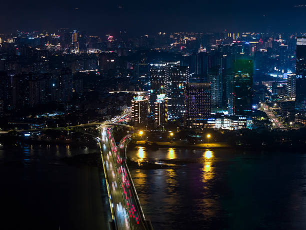A suspension-style bridge bathed in vivid blue lighting spans a calm river at night. String lights outline the bridge’s cables and towers, creating a near-perfect mirrored reflection on the water. Warm-toned city buildings are visible in the background, offering a striking color contrast ideal for urban night photography and architectural lighting showcases