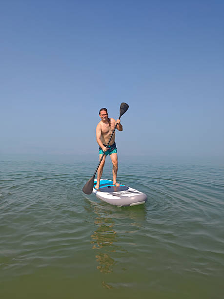A serene summer view of Lake Balaton's calm, light blue waters with a lone person wading in the distance, framed by a vibrant green lawn and scattered rocks in the foreground under a hazy sky.