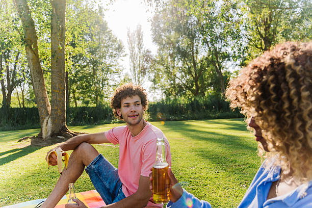 Young multi-ethnic couple sitting on a blanket in a park, enjoying a delightful picnic with drinks on a sunny summer day, radiating happiness and carefree love