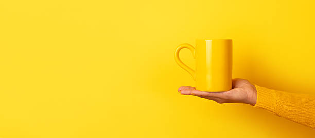 Hand in yellow sweater presenting a yellow ceramic mug on open palm against a solid yellow background.