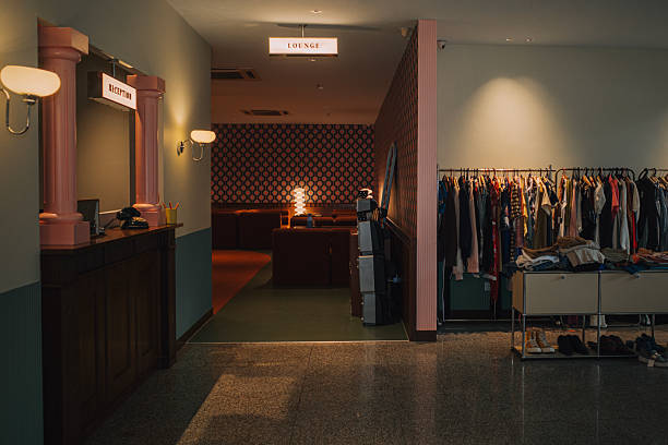 A stylish boutique hotel lobby interior featuring a retro-themed reception counter with pink columns, vintage telephone, and a cozy lounge in the background. A curated clothing rack and soft warm lighting complete the nostalgic yet modern aesthetic. Ideal for themes related to travel, lifestyle, interior design, or hospitality concepts.