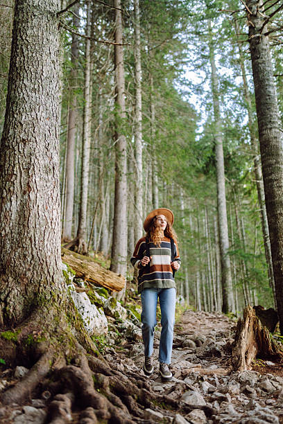 A young woman with a backpack and a hat walks along a rocky path in a dense forest. A female traveler enjoys nature and hiking. Travel, hiking and nature concept. Active lifestyle.