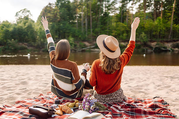Beautiful girlfriends relaxing on blanket by river and enjoying hot drinks, reading a book at sunset. Two young women having fun and spending time together outdoors. Friendship, relaxation concept.