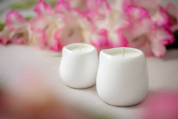 White ceramic candles on a soft surface with blurred pink flowers in the background. Minimalist spa composition for relaxation, wellness, aromatherapy, and home decor concepts.