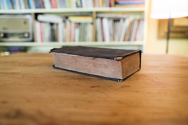 An Old and Worn Bible With Frayed Binding and Faded Pages Resting on a Wooden Table, Set Against a Softly Blurred Background of a Cozy Home Library With Bookshelves. The Image Evokes Faith, Tradition, and the Passage of Time.