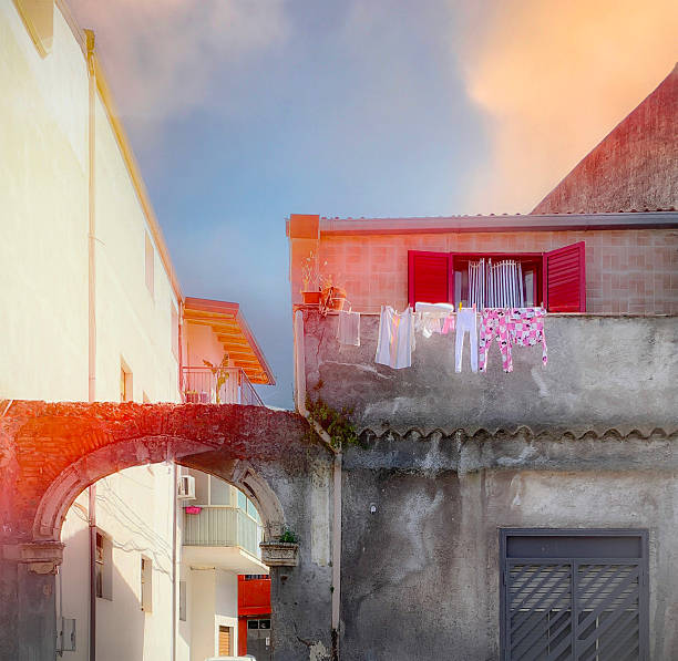 Authentic Mediterranean street view in Catania, Sicily. A clothesline with colorful laundry stretches across the front of a weathered building with red shutters and textured walls. An old stone arch connects two adjacent buildings, under a warm and soft afternoon light.
