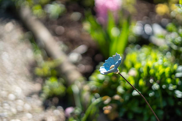A charming, close-up photograph of a small, light blue ceramic flower ornament with a shallow depth of field. The handmade decoration stands out on its thin stem against a beautifully blurred background of a sunny garden, with hints of green foliage and a stone path. The focus is sharp on the delicate details of the ceramic petals, while the soft bokeh adds a peaceful and whimsical atmosphere. This image is perfect for themes of gardening, decoration, craftsmanship, home and garden, and creative hobbies.