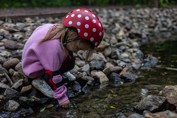 A curious child in protective gear, including a red helmet with white dots and elbow pads, explores nature by touching the cool water of a shallow stream. A child explores a small rocky stream..