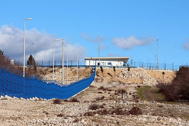 A high blue security fence topped with spikes leads to a modern building on a hill, surrounded by tall lighting poles and rough, rocky terrain under a bright sky