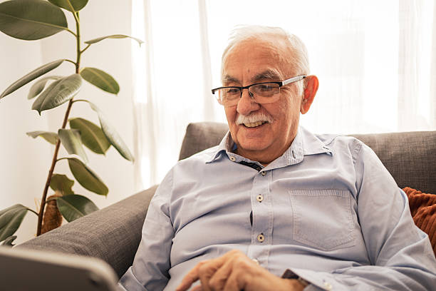istock Senior man smiling while using a tablet on a cozy living room sofa 2227948081